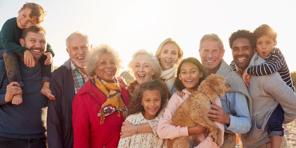 11 extended family members of various ages posing for a group portrait with the sun at their back. One of the children standing in the front row is holding a small golden-brown dog. 2 children are being held on their back. 